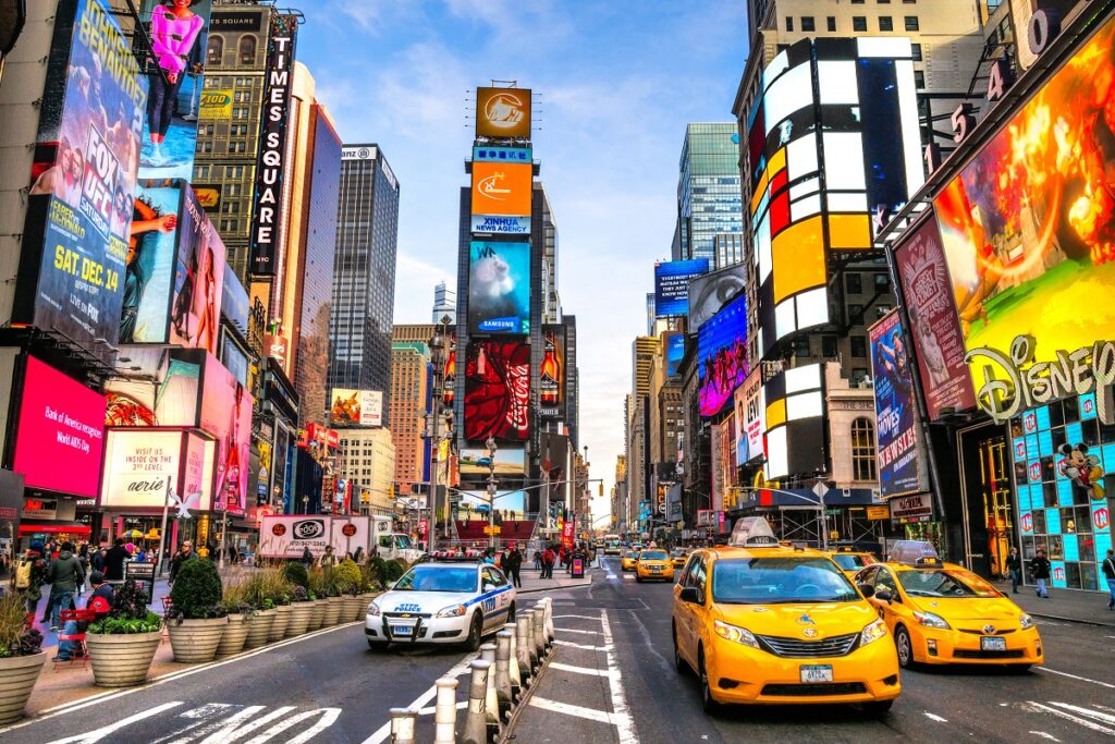 Imagem de Times Square em Nova Iorque, com seus icônicos painéis luminosos, multidões de pessoas e táxis amarelos. Times Square é um movimentado centro comercial e de entretenimento conhecido por seus teatros, lojas e anúncios brilhantes. Saiba mais!